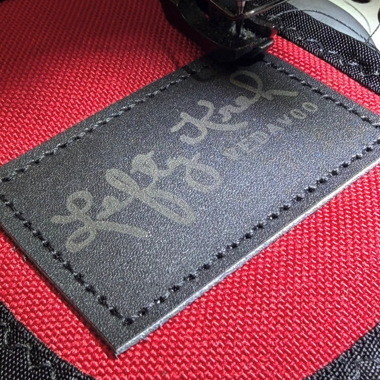A close-up of a black leather patch with engraved signature of Lefty Kreh and the text 'VEDAVOO' on a red and black Cordura fabric, with a sewing machine needle visible at the top.